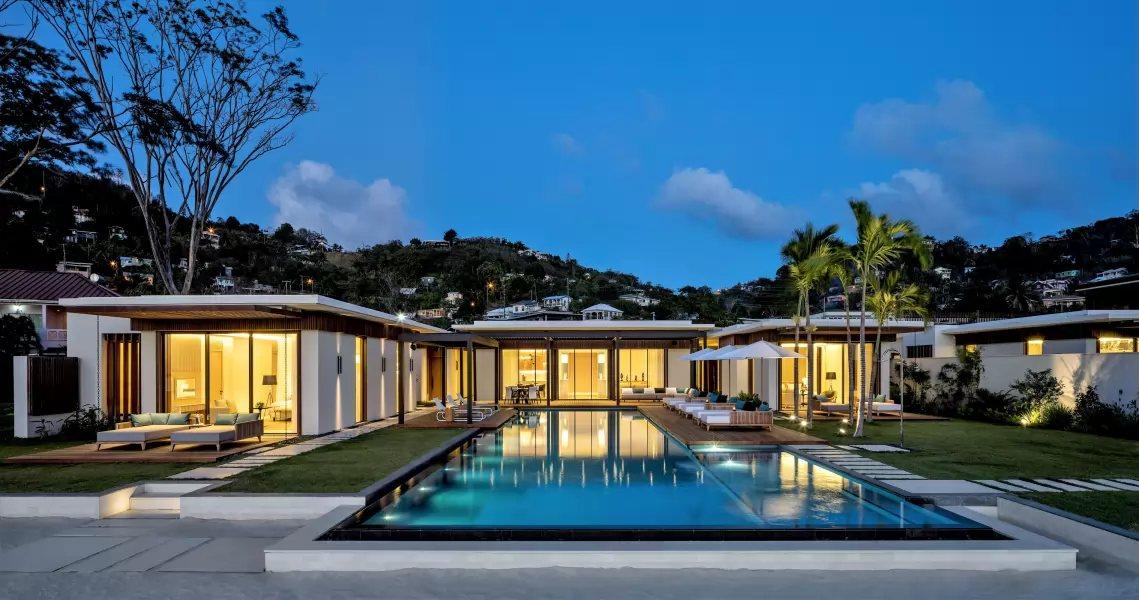A modern single-storey villa with a pool and lawn at dusk, with houses on the hillside behind